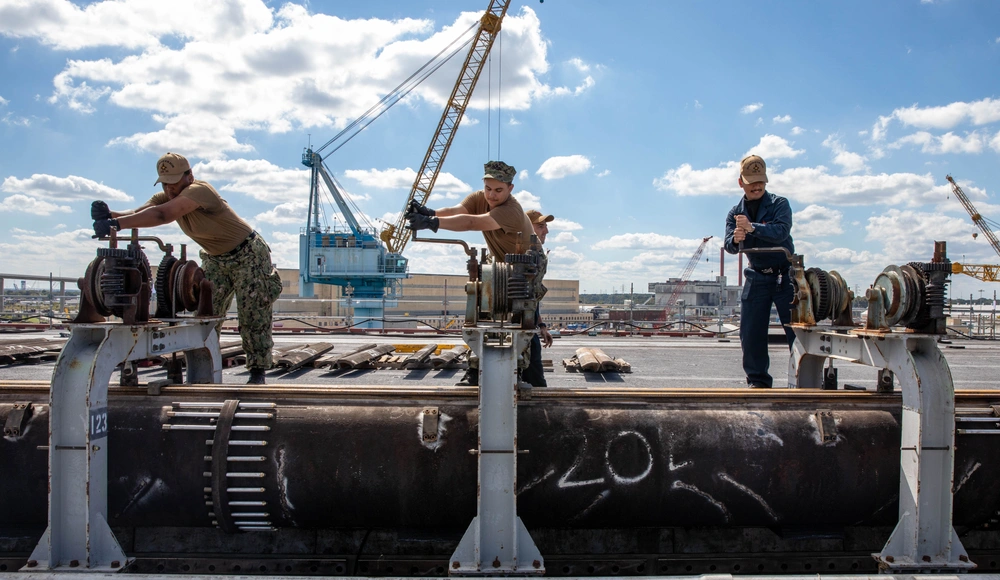 Sailors hand-crank a large steam-catapult cylinder during maintenance on USS George H.W. Bush in 2024.